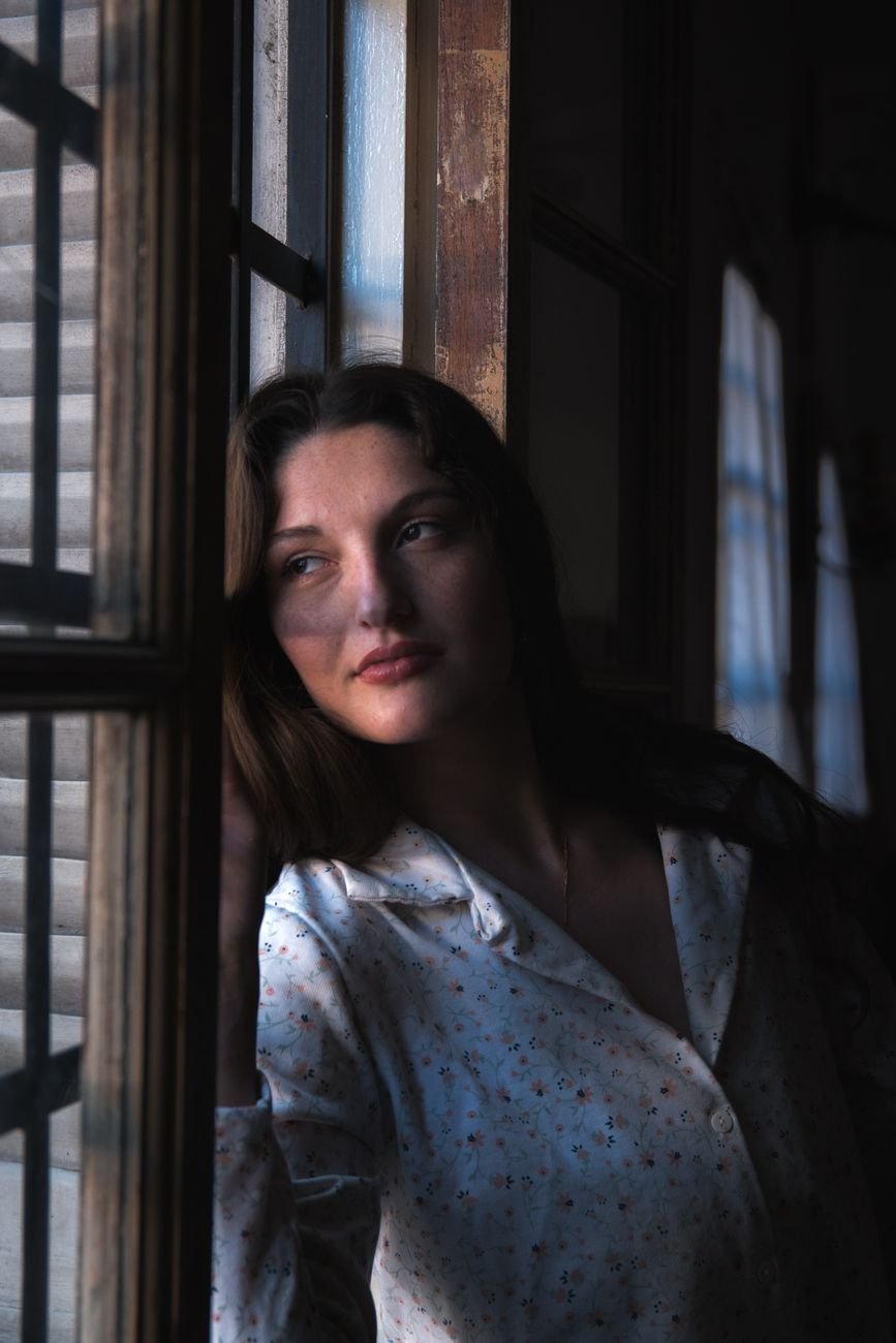 Kobieta w zamyśleniu przy oknie, miękkie światło — spokój i refleksja